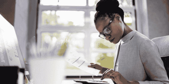 Mulher sentada, em frente à uma mesa, utilizando uma calculadora e um computador, enquanto segura documentos, fazendo cálculos.