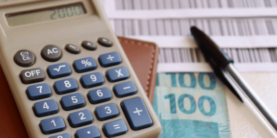 Mesa de escritório com calculadora, dinheiro de papel, caneta e boletos, uma forma de parcelamento recorrente.